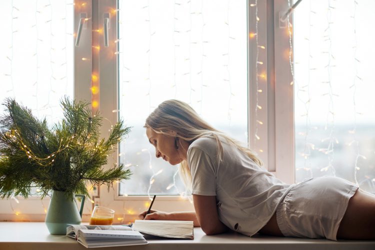 Beautiful young woman in the Christmas holidays reading a book at the window of the house